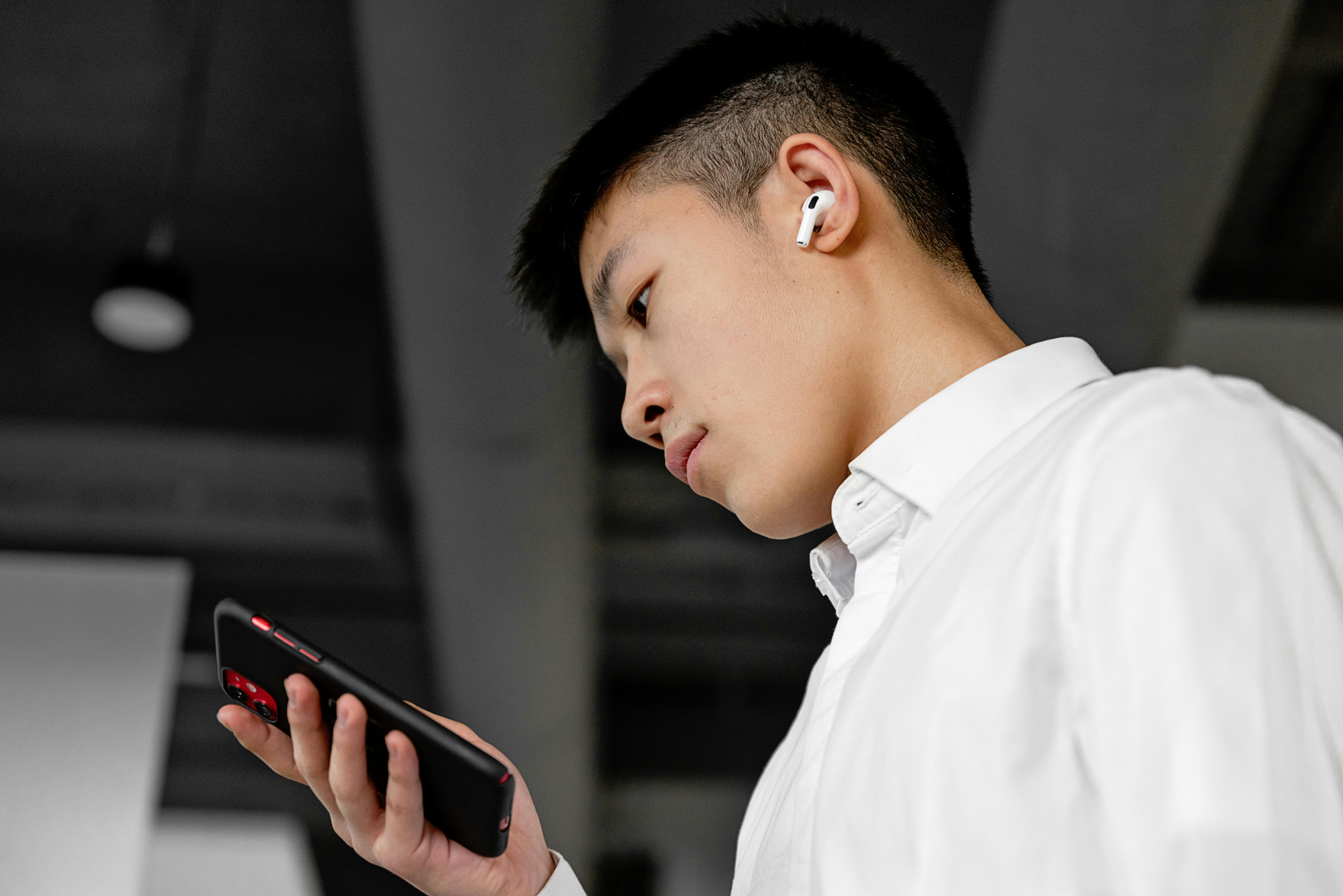 Side view of a young man wearing wireless earbuds and using a smartphone indoors.